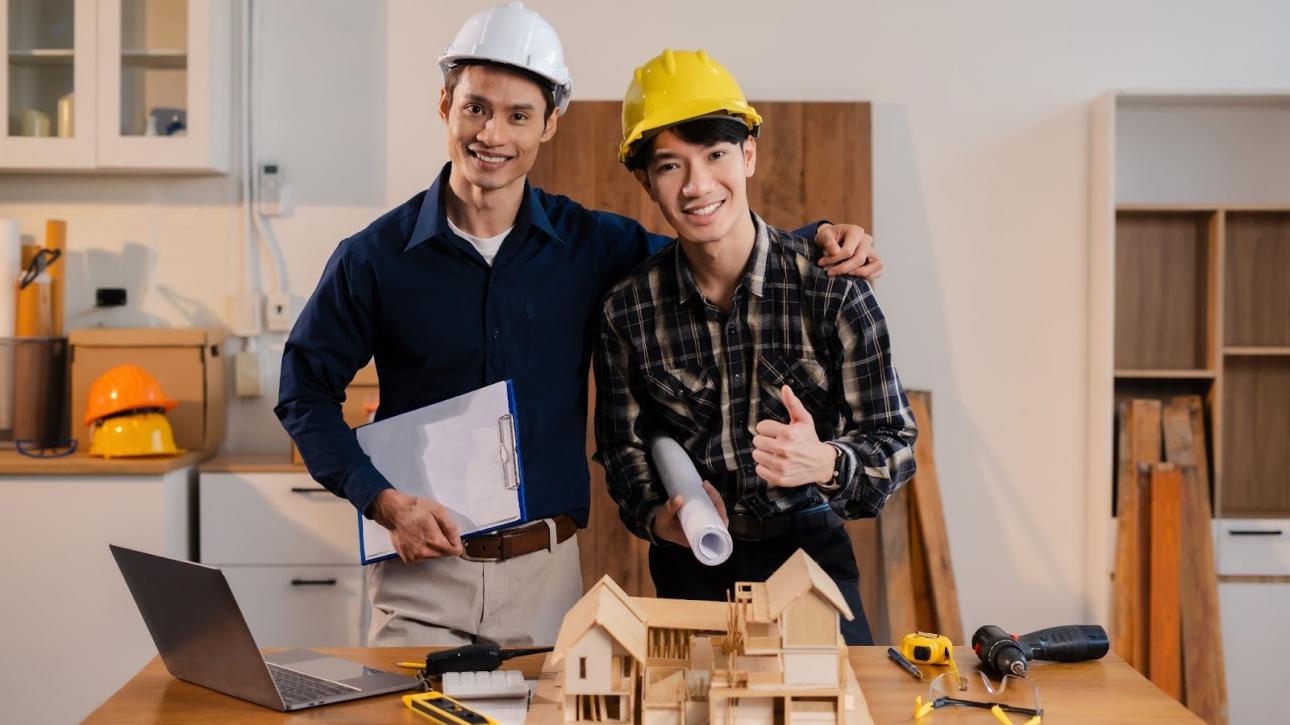 Home remodeling contractors at a desk with a model house, tools, a laptop, and blueprints; one gives a thumbs-up while the other holds a clipboard