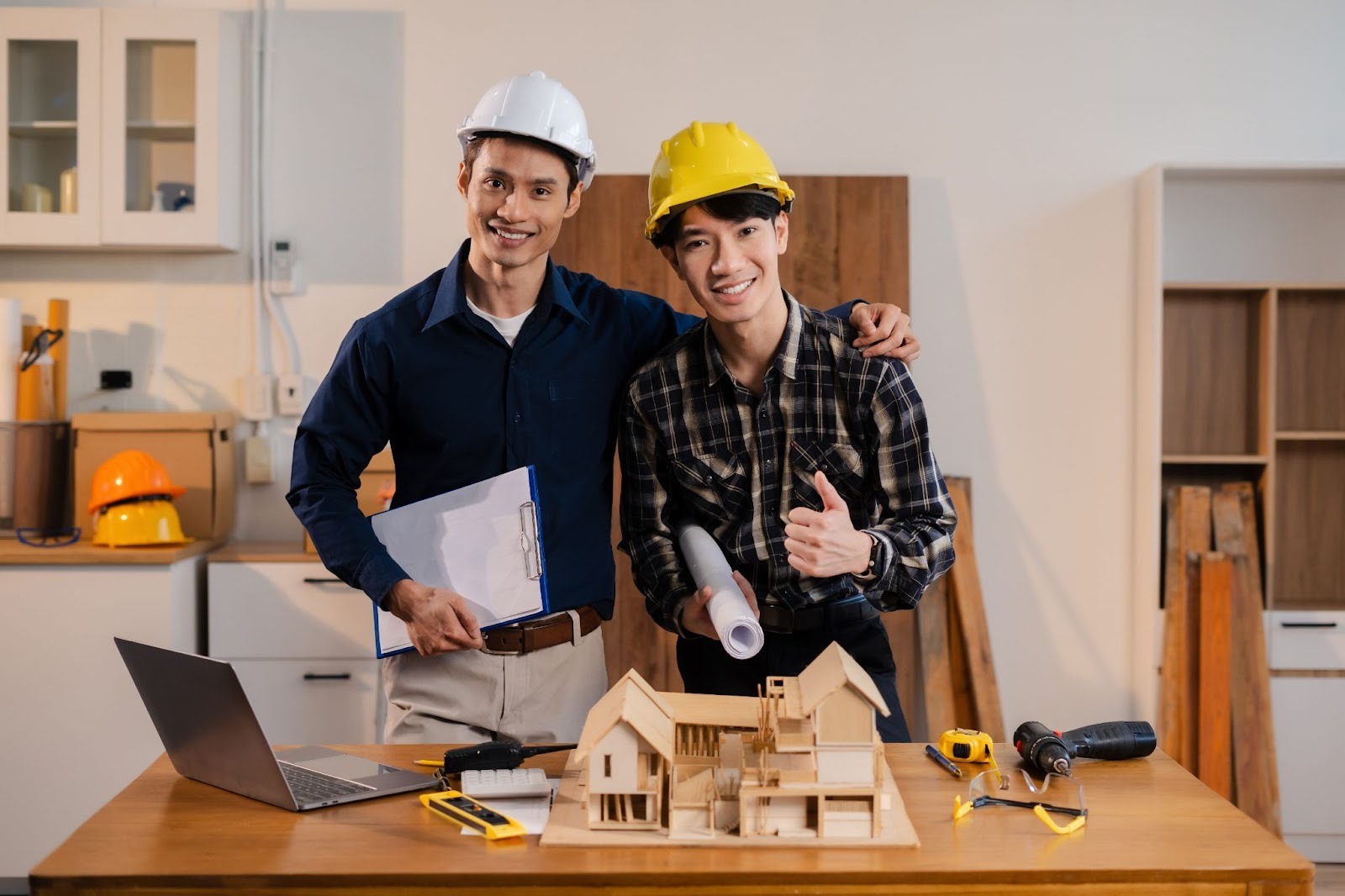 Home remodeling contractors at a desk with a model house, tools, a laptop, and blueprints; one gives a thumbs-up while the other holds a clipboard