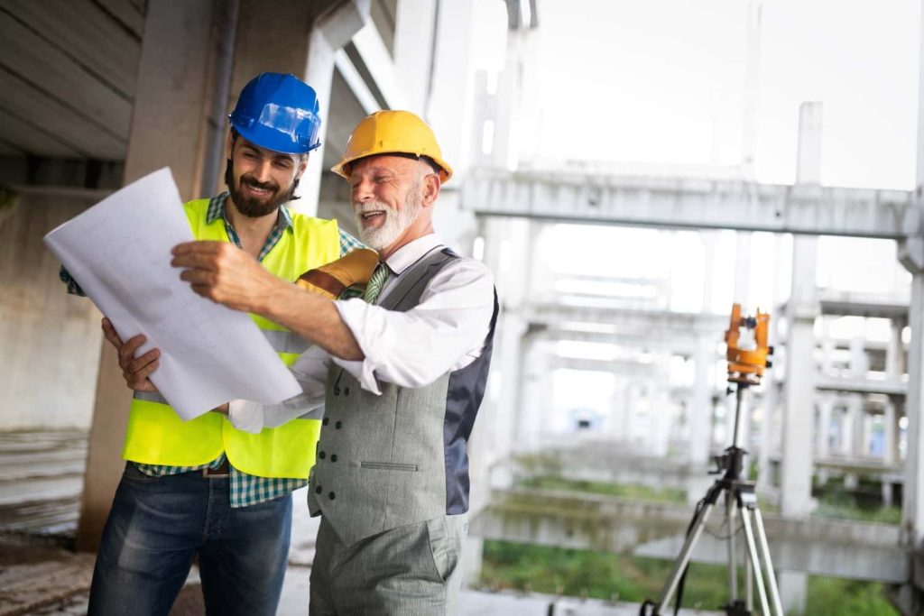 An older man in a yellow hard hat and gray vest points at a large blueprint with a younger man in a blue hard hat and safety vest, in an industrial construction site.