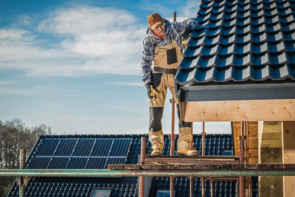 A construction worker in a plaid shirt and tan overalls stands on scaffolding, inspecting the dark, wavy roof tiles of a house under a blue sky.
